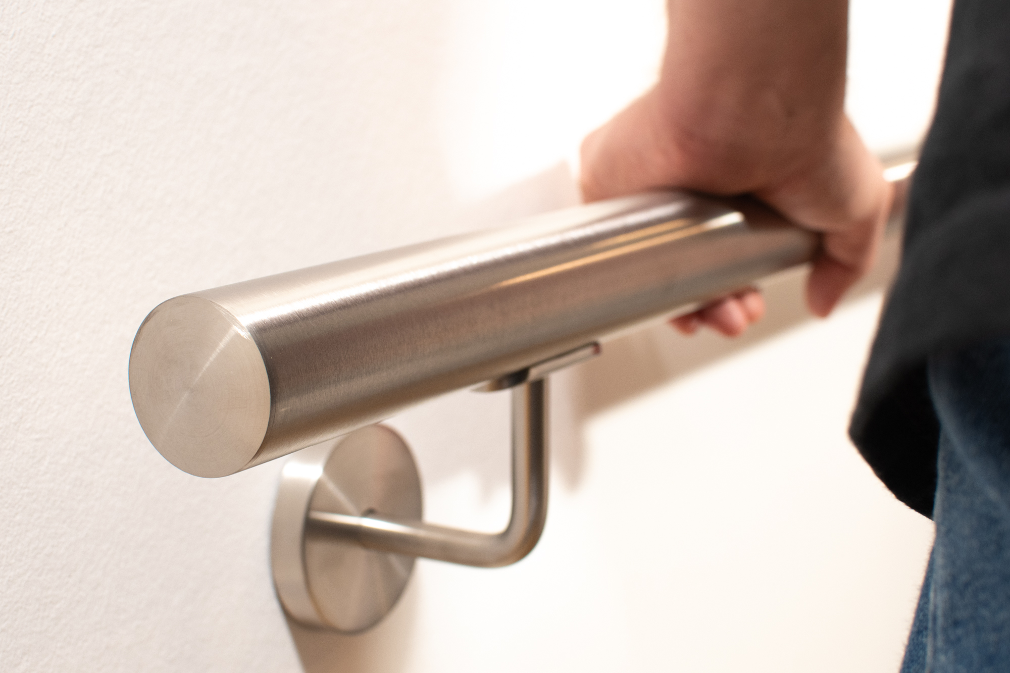 Main courante ronde en acier inoxydable sur mur blanc avec support inox, une main la saisit, about plat au premier plan