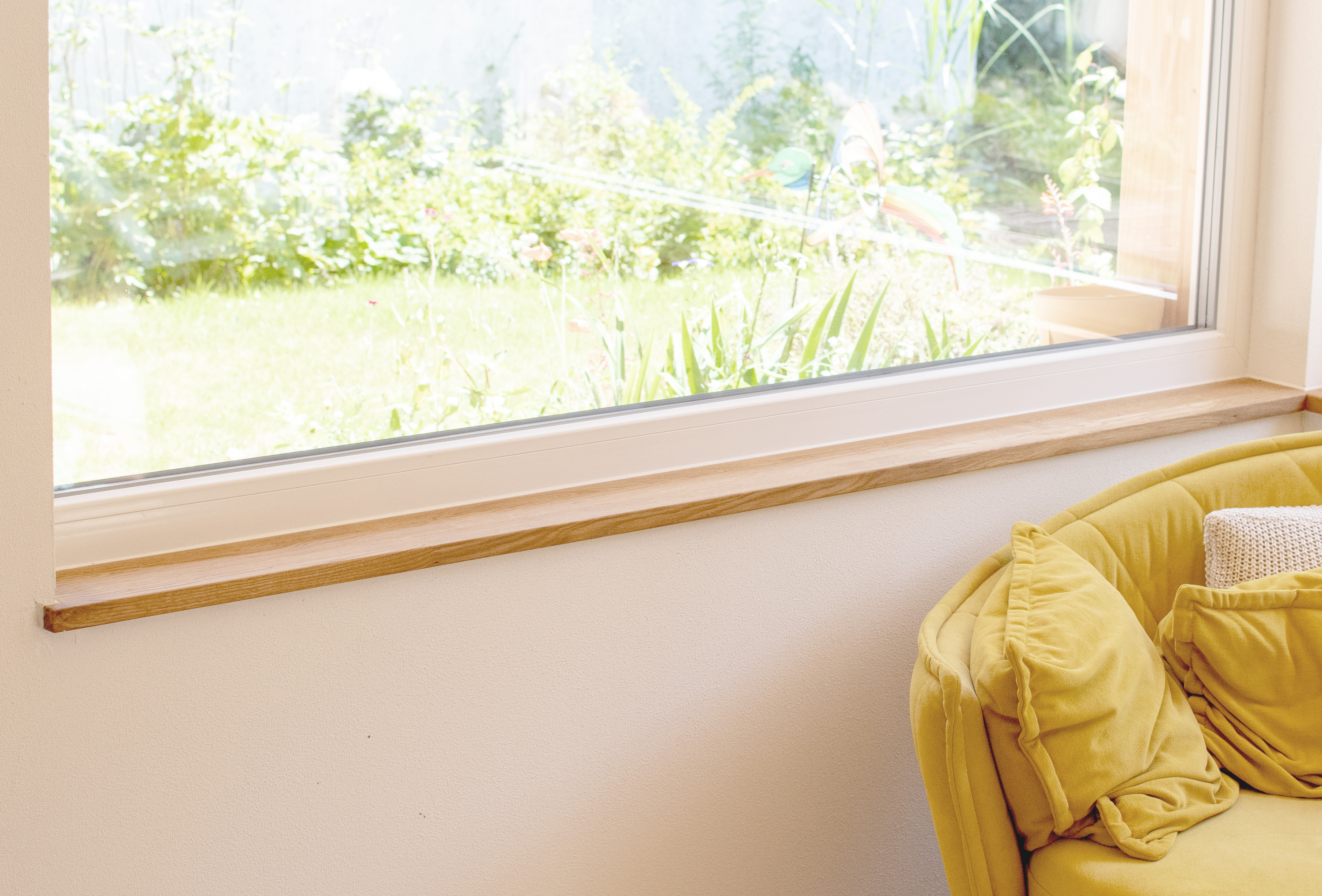 Appui de fenêtre étroit en bois clair sous une large fenêtre donnant sur le jardin, canapé jaune avec coussins au salon