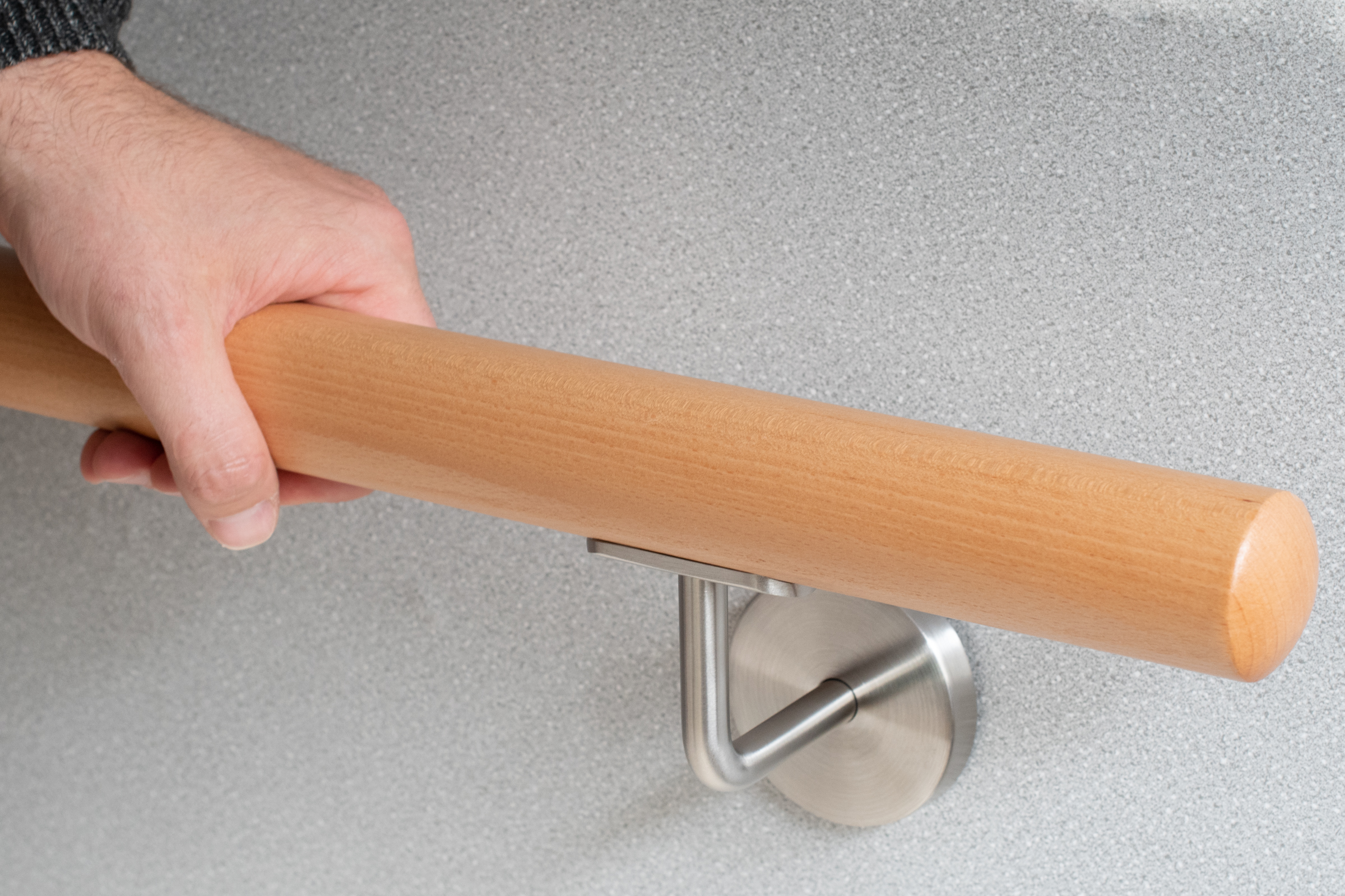 Main courante ronde en hêtre sur mur gris avec support inox, une main saisit la main courante, détail de la prise