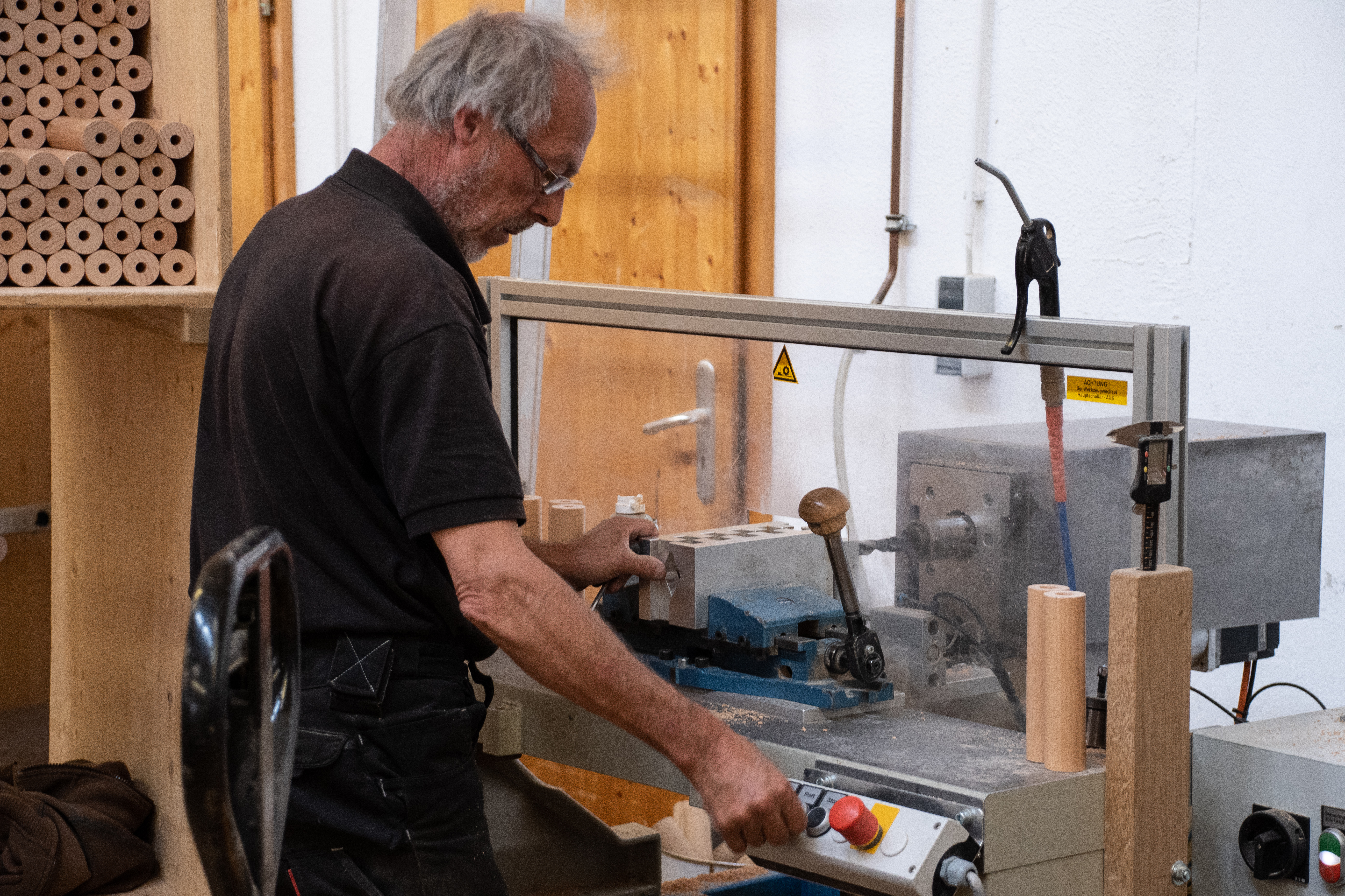Fabrication de mains courantes : employé perçant une pièce en bois à la perceuse à l'atelier, étagère de bois ronds derrière