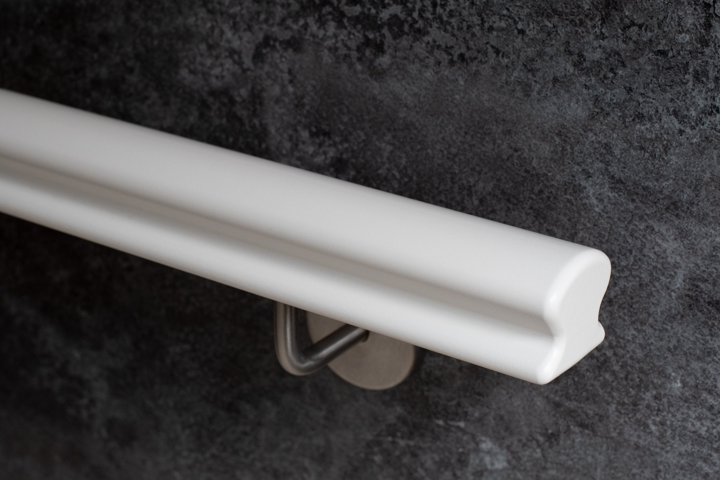 Main courante en bois Omega laquée blanc avec support mural inox devant un mur gris foncé, gros plan du profil