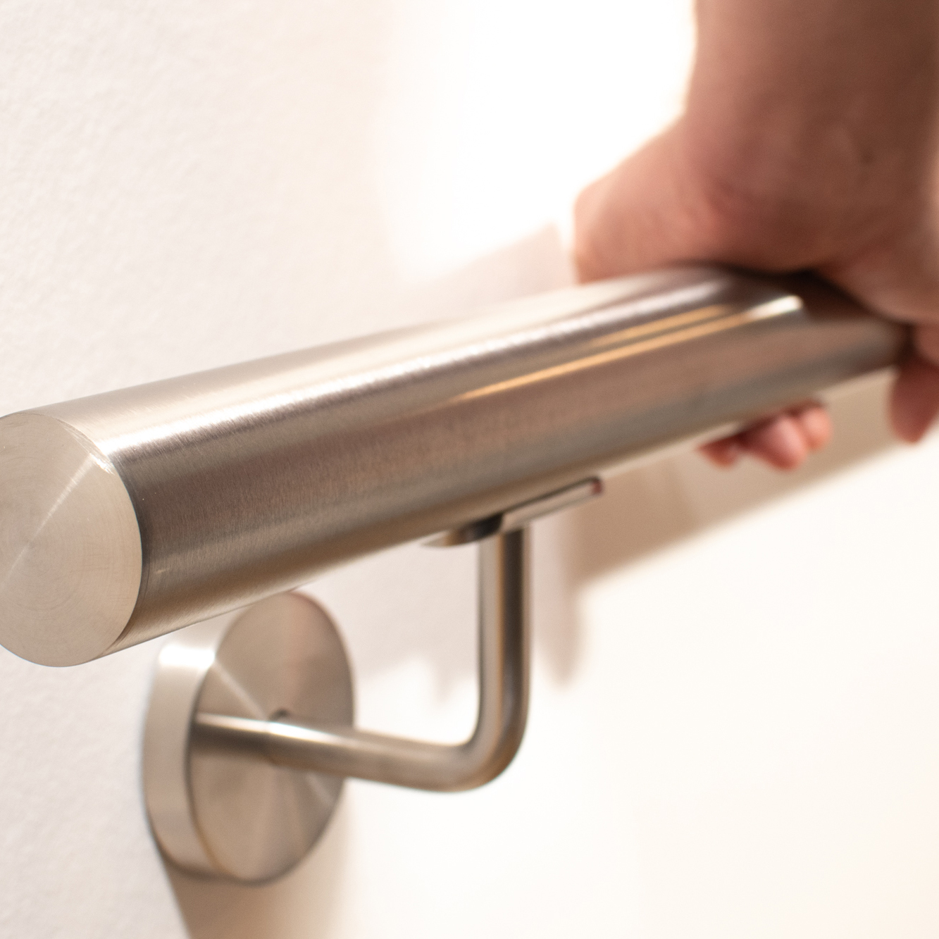 Main courante ronde en acier inoxydable au mur blanc avec support mural inox, une main saisit le tube brossé