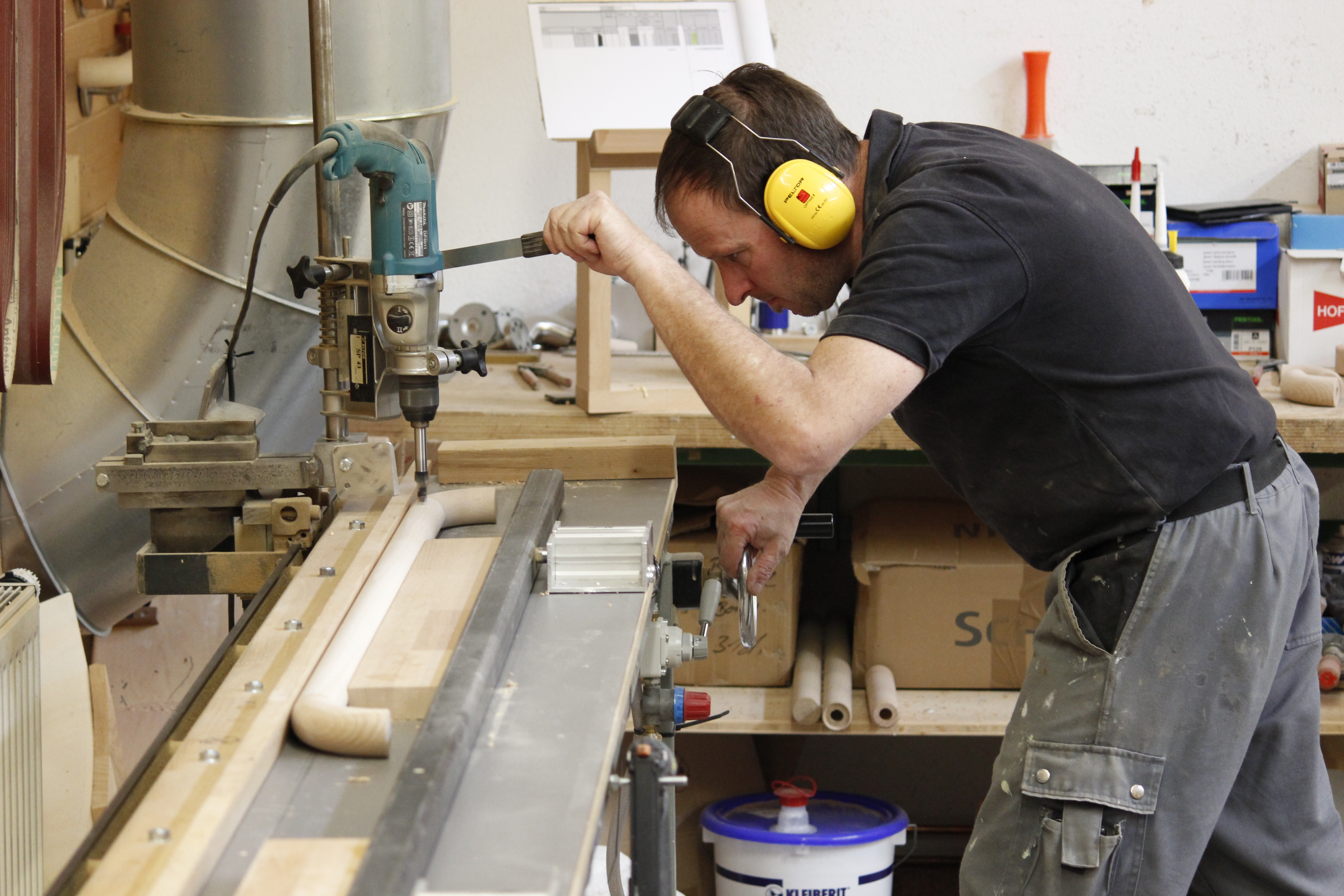 Artisan avec protection auditive perçant des trous dans une main courante en bois sur une perceuse à colonne à l'atelier