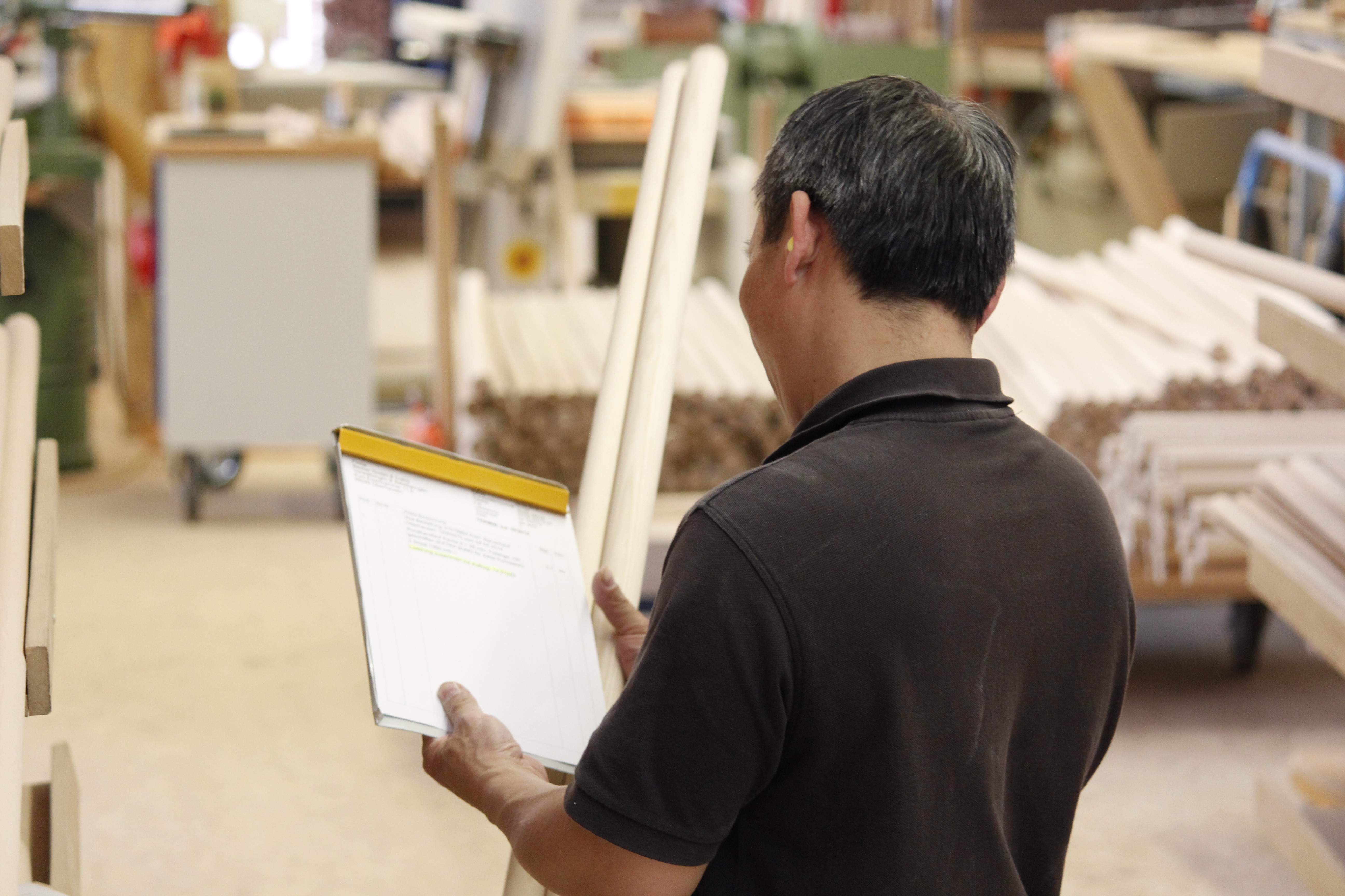 Employé avec dossier de commande dans l'atelier bois, barres rondes et ébauches de mains courantes derrière, vu de dos