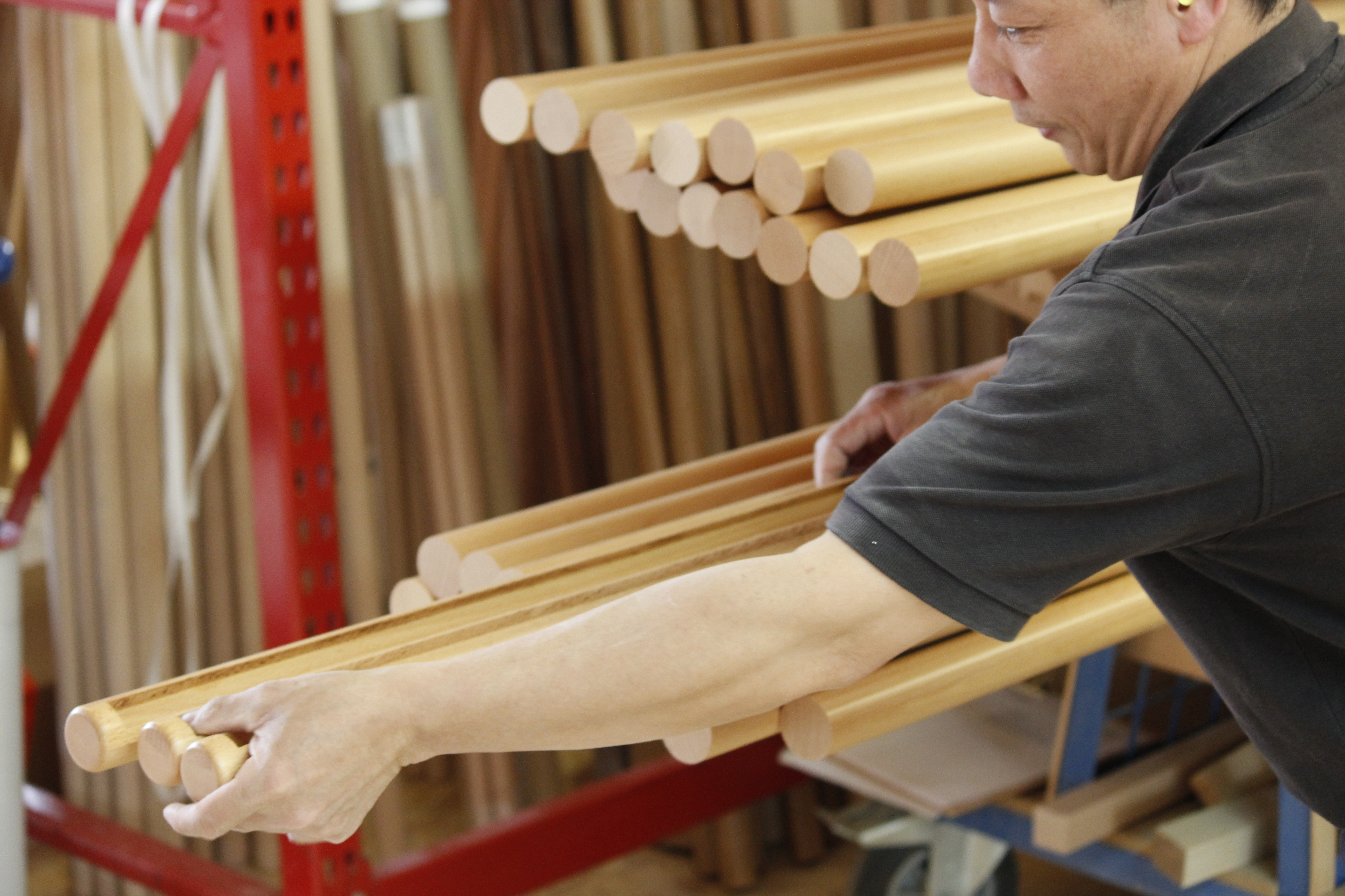 Mains courantes rondes en hêtre portées en fagot par un employé à travers l'atelier, étagère de barres en bois derrière