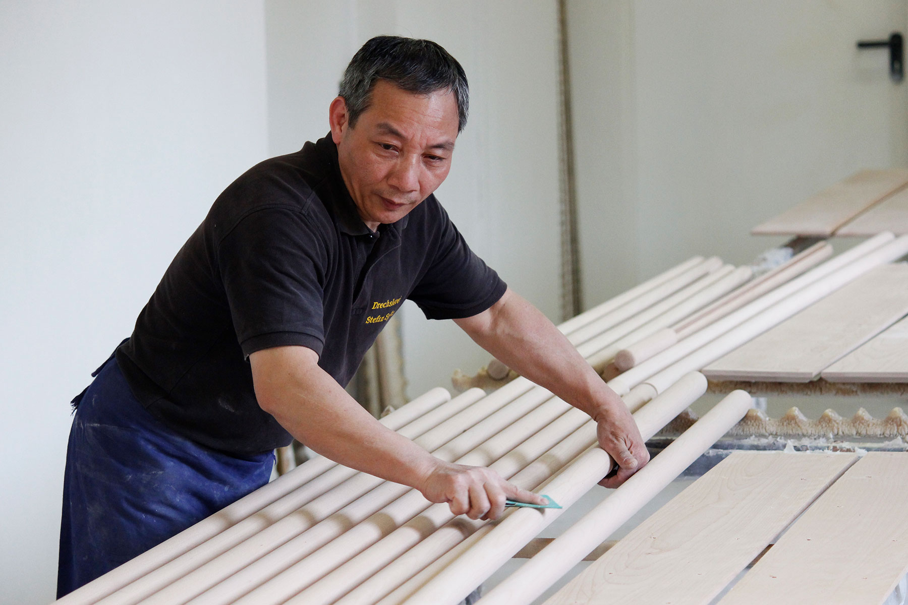 Fabrication de mains courantes : employé contrôlant des barres rondes en bois clair à l'atelier, planches en arrière-plan